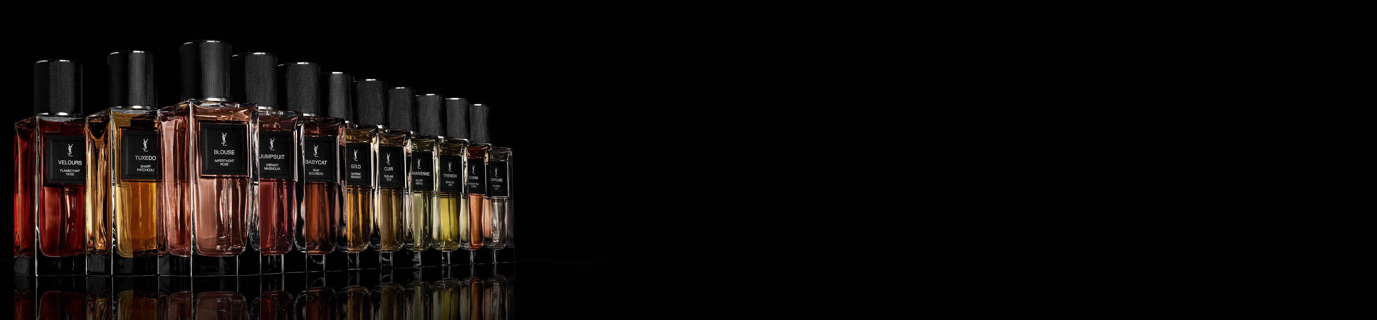 Una fila di flaconi di profumo Yves Saint Laurent con liquidi di vari colori e tappi neri testurizzati, che si riflettono su una superficie scura su sfondo nero.