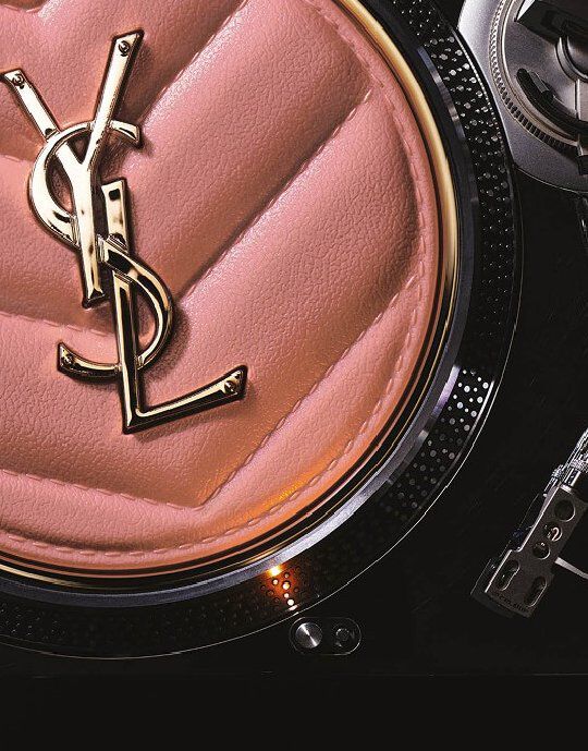 Primo piano di una pochette o accessorio Yves Saint Laurent rosa trapuntato con il logo YSL dorato al centro. L'oggetto e posizionato sul bordo metallico e testurizzato di un giradischi, fondendo il lusso della bellezza con un'estetica ispirata alla musica.