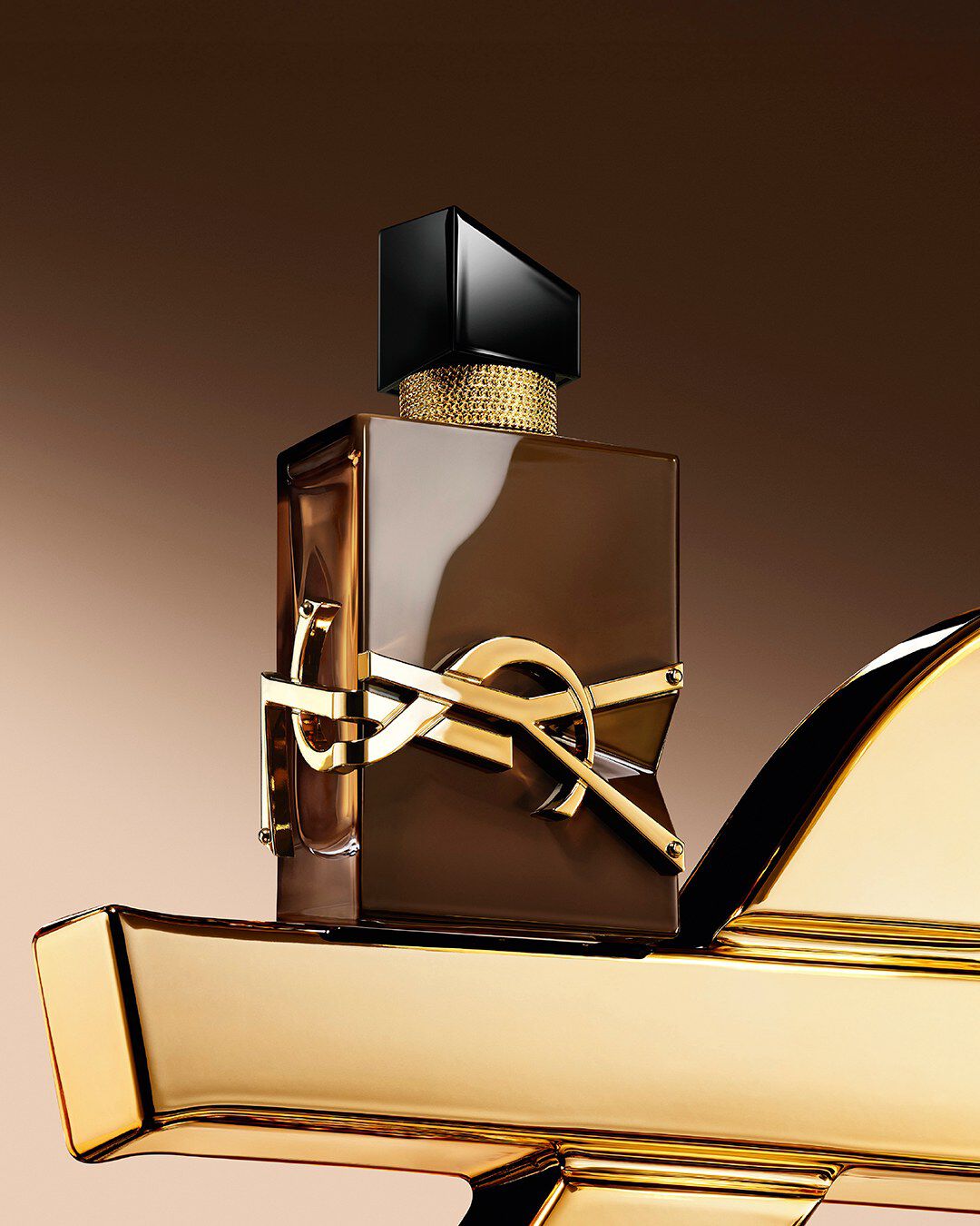 Flacone Libre Santal Couture marrone smaltato che riflette la luce, posato su una grande Cassandre YSL dorata.