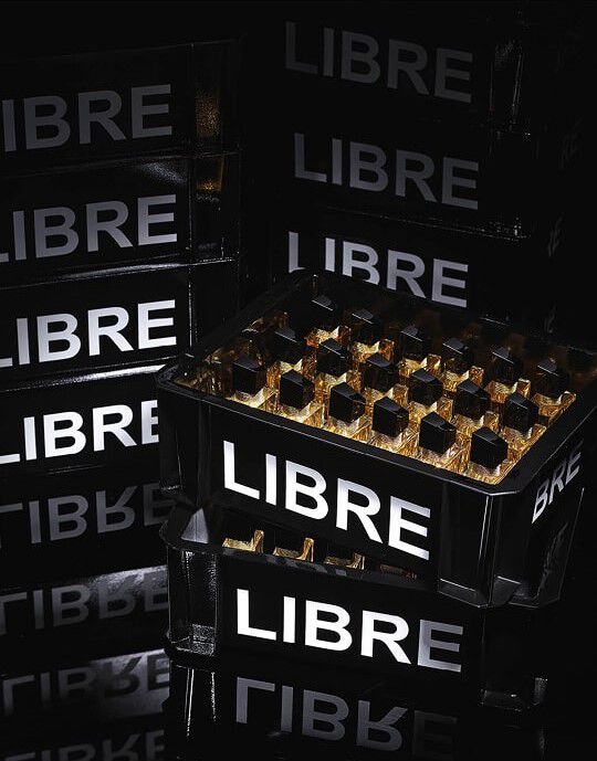 Una composizione di casse nere, simili a quelle utilizzate per il trasporto di bottiglie, impilate e riempite di flaconi del profumo YSL Libre. Le casse recano l'iscrizione "LIBRE" in grandi lettere bianche luminose. In primo piano, una cassa e piena di numerosi flaconi del profumo "Libre" che riflettono la luce, creando bagliori dorati. L'intera scena e immersa in una luce fioca, accentuando il lato chic e la messa in scena del prodotto.