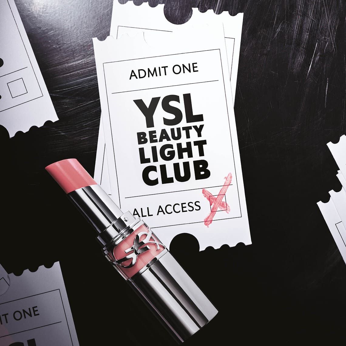 Un rossetto rosa chiaro dalla texture lucida e posato su due biglietti bianchi di tipo evento, leggermente sovrapposti. Il biglietto superiore reca la scritta "ADMIT ONE" in alto, e al centro, "YSL BEAUTY LIGHT CLUB" in grandi lettere nere, con "ALL ACCESS" sotto. Una croce rossa e scarabocchiata nell'angolo inferiore destro del biglietto, come per convalidarlo. Lo sfondo e scuro, evocando una superficie testurizzata, e si intravedono i bordi strappati di altri biglietti sui lati, suggerendo la presenza di diversi altri tagliandi sparsi. L'insieme da l'impressione di un invito esclusivo a un evento glamour.
