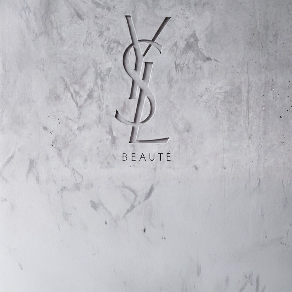 Logo YSL BEAUTE inciso su una parete in cemento grigio strutturato.