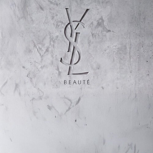 Logo YSL BEAUTE inciso su una parete in cemento grigio strutturato.