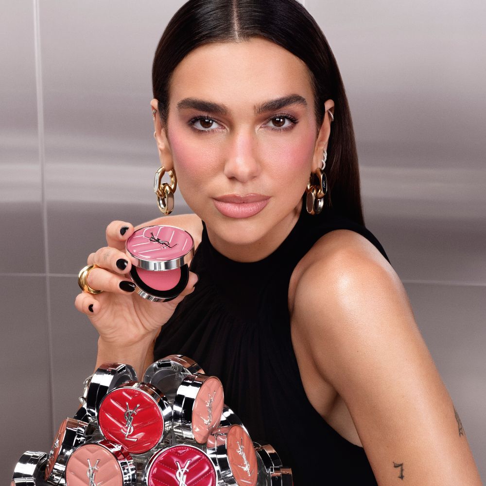 Dua Lipa tiene in mano il compact aperto di YSL Make Me Blush Lifted Glow Cream Blush, con una collezione delle nuove tonalita esposte davanti a lei.