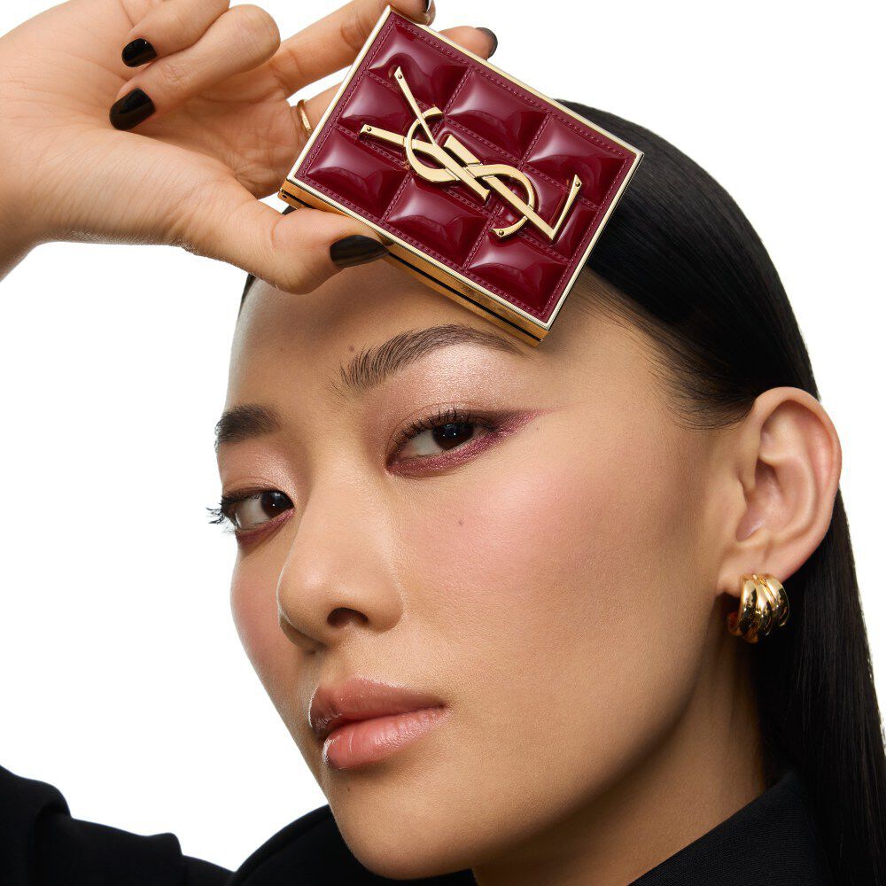 Modella in completo nero e look glamour, seduta in un’atmosfera festiva illuminata dalle candele, circondata da cofanetti regalo YSL.