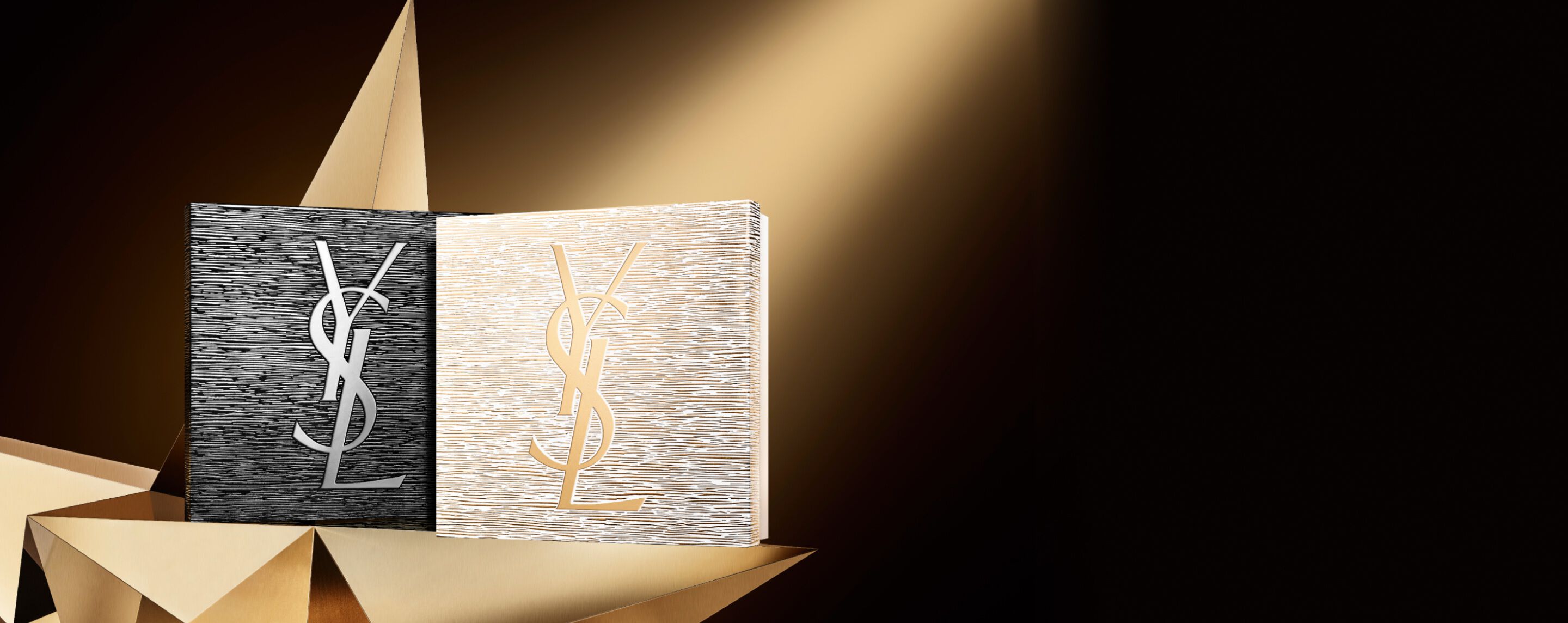 Due eleganti cofanetti regalo natalizi YSL Beauté, uno nero con logo argentato e uno dorato con logo dorato, posizionati su una superficie geometrica dorata, che allude all'alta moda e alla brillantezza.