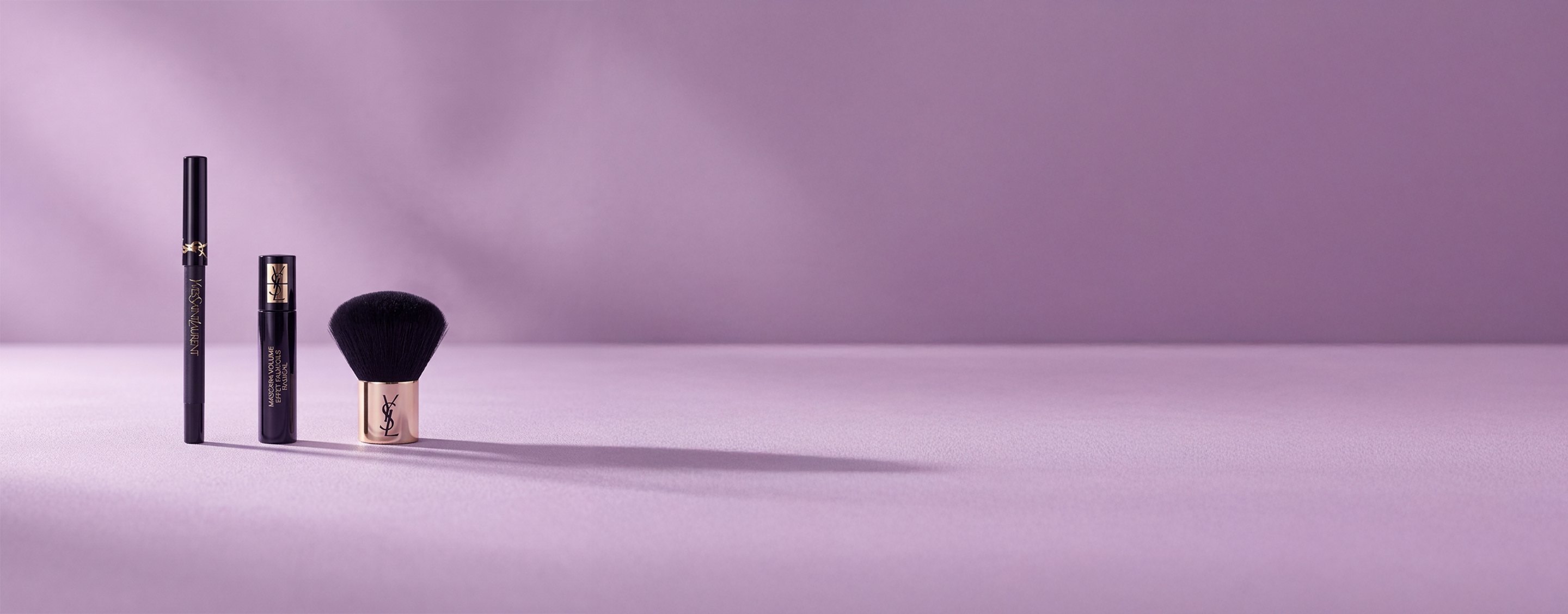 Tre prodotti di bellezza Yves Saint Laurent, tra cui un mascara e un pennello kabuki, su un morbido sfondo color lavanda.