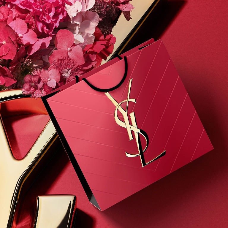 Confezione regalo YSL rosa acceso con logo Cassandre dorato, circondata da petali di fiori primaverili.