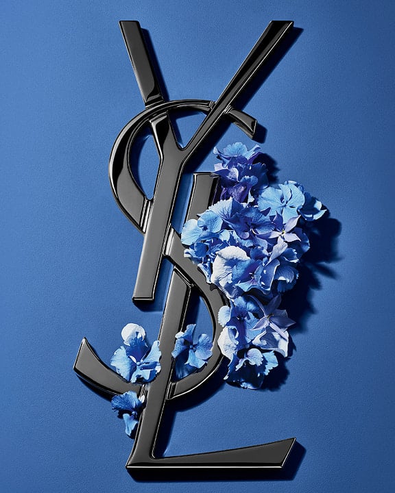 Logo Cassandre YSL nero ornato da vivaci fiori primaverili blu, su uno sfondo blu sereno.