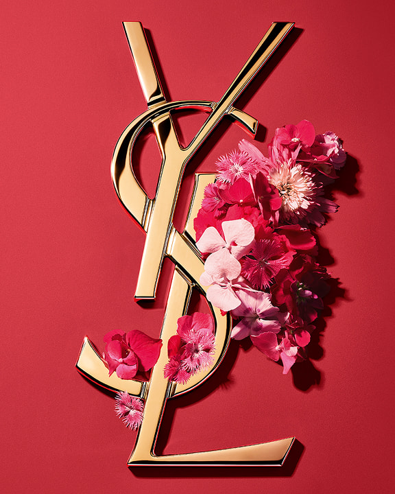 Logo Cassandre dorato YSL, ornato con vibranti fiori primaverili rosa, su uno sfondo rosa.