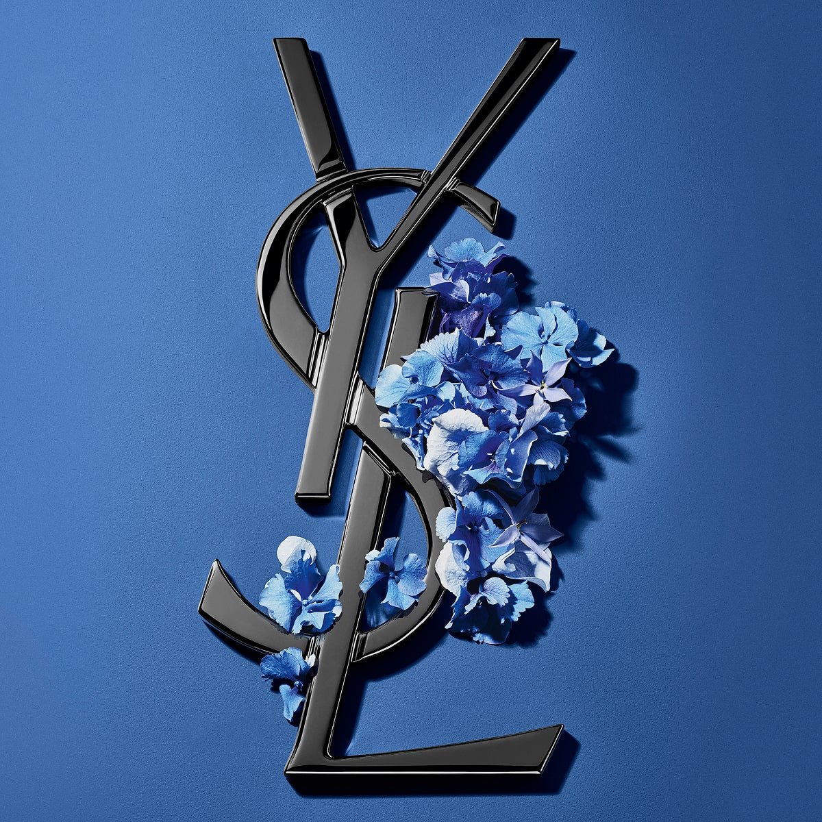 Logo Cassandre YSL nero ornato da vivaci fiori primaverili blu, su uno sfondo blu sereno.