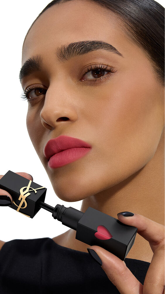 Modella con tono di pelle medio, labbra in un rosa-mat vibrante, tiene il flacone nero di YSL THE INKS BLUR con il cuore rosa.