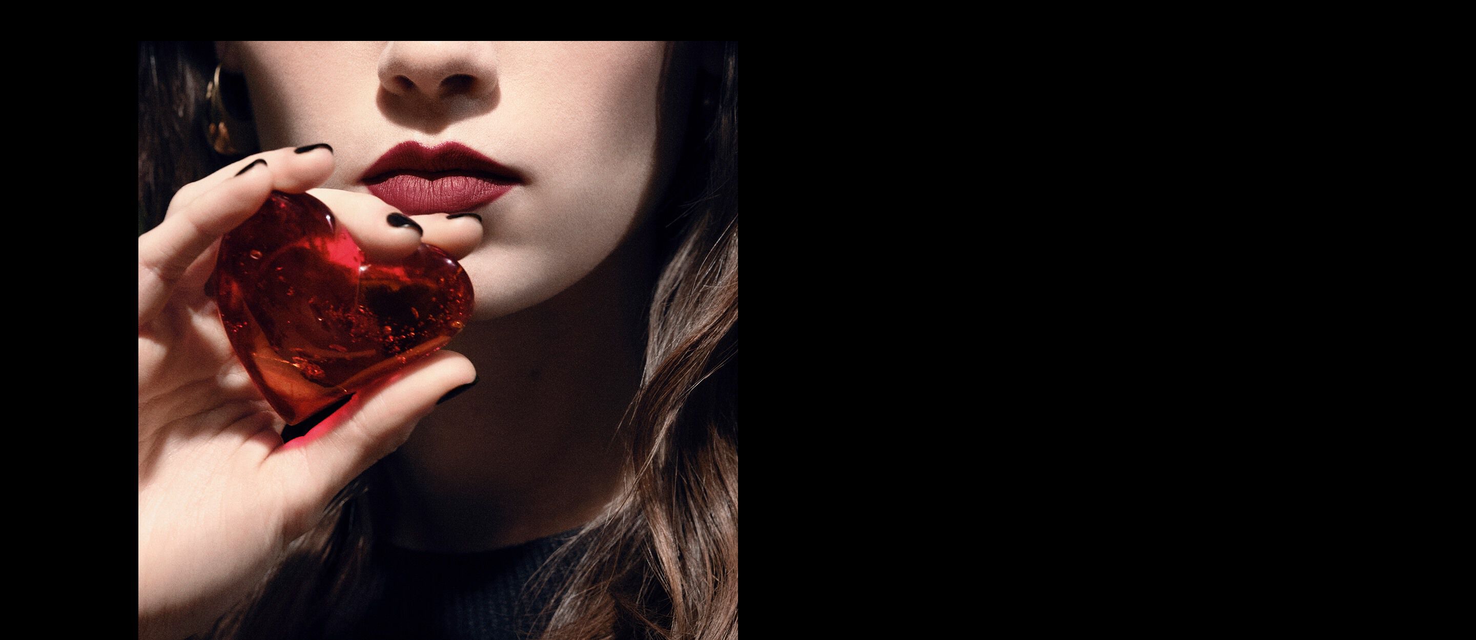 Primo piano delle labbra e della mano di una modella
che tiene un gioiello a cuore rosso rubino
traslucido per YSL CŒUR Eau de Parfum.