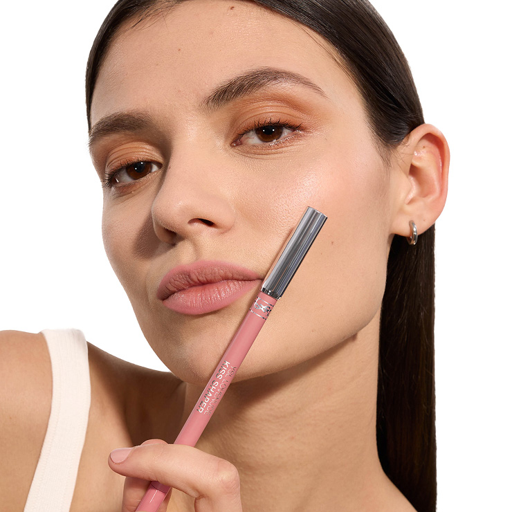 Primo piano di una modella con labbra opache rosa tenue che tiene una matita labbra YSL Kiss Shaper coordinata.