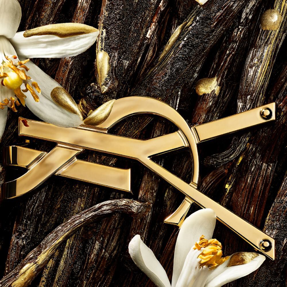 Primo piano del logo YSL dorato posizionato tra baccelli di vaniglia, fiori bianchi e accenti dorati scintillanti.