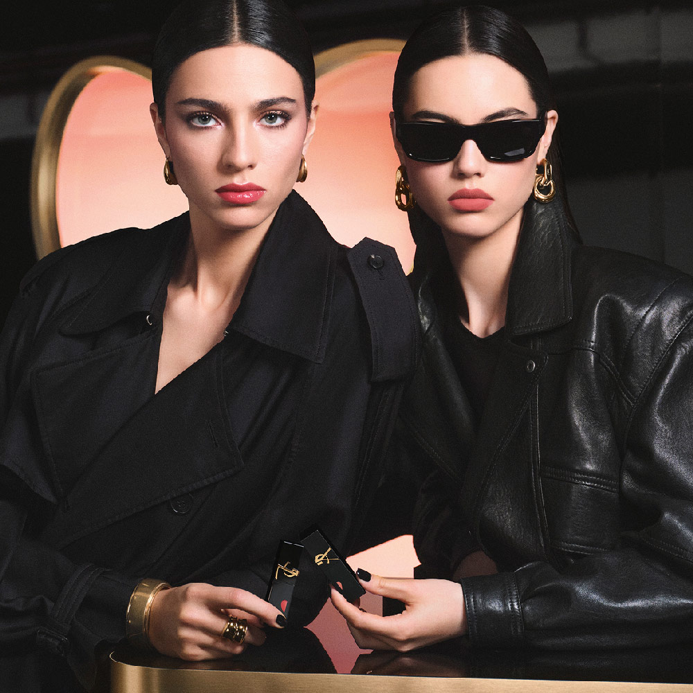 Due modelle chic in trench nero e giacca di pelle reggono rossetti YSL davanti a uno sfondo rosa a forma di cuore, mostrando un look labbra lucide rosse e un effetto matte rosa delicato.
