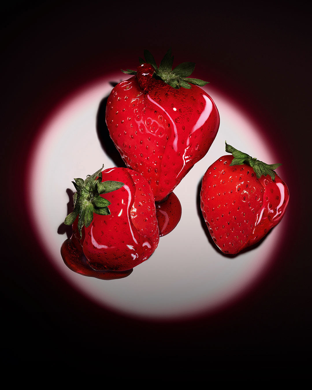 Tre fresche fragole rosse illuminate da un riflettore rosa circolare su uno sfondo scuro.