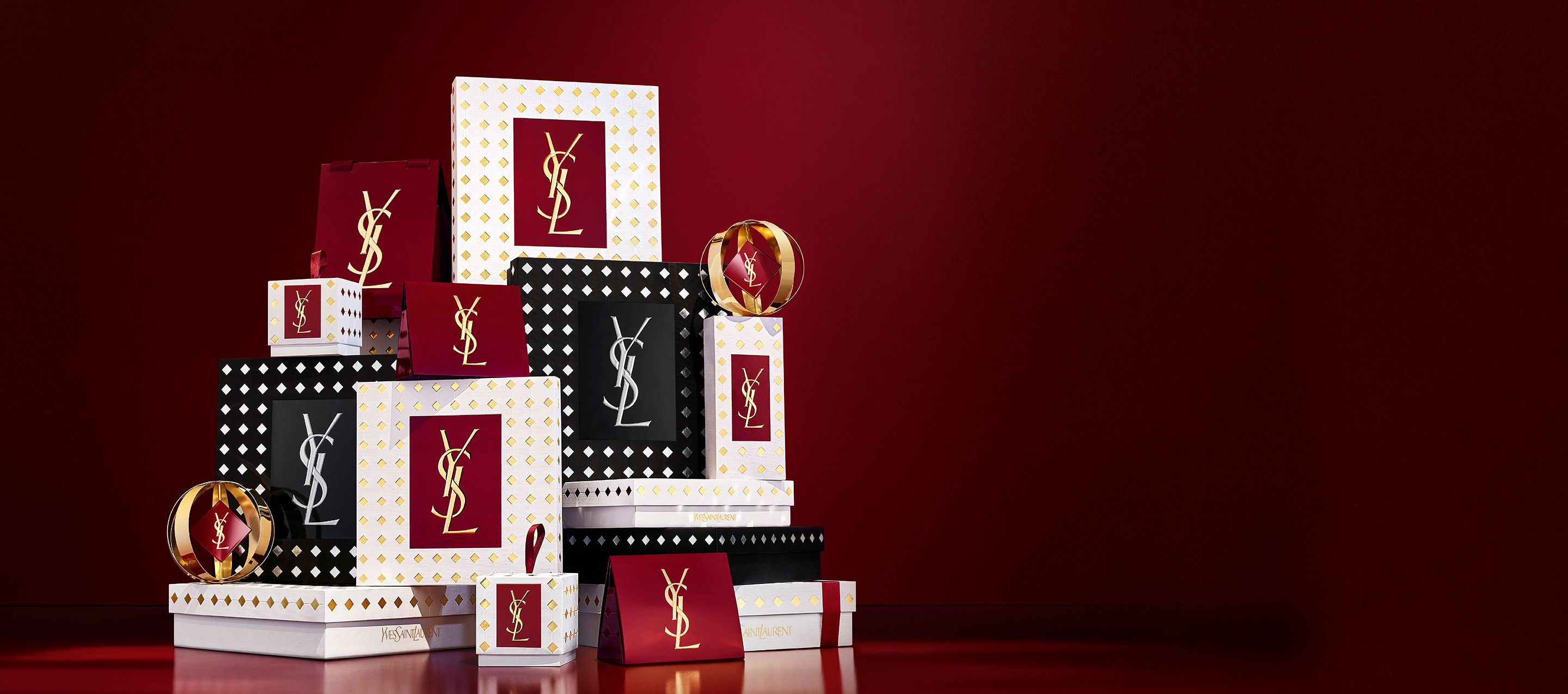 Tre modelle con eleganti outfit neri tengono cofanetti regalo YSL davanti a un sipario rosso festivo con coriandoli dorati che cadono.