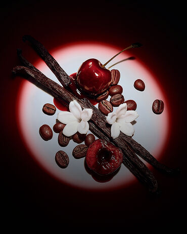 Primo piano di succose ciliegie rosse, fiori di gelsomino, bacche di vaniglia e chicchi di caffè illuminati da un drammatico fascio di luce per Black Opium Eau de Parfum Over Red.