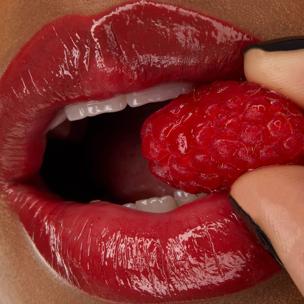 Primo piano delle labbra della modella Zahara, impreziosite dal rossetto YSL LOVESHINE nella tonalità Berry Crush 219, mentre tiene tra i denti una lampone fresco.