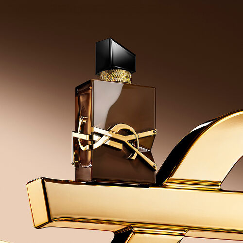 Flacone Libre Santal Couture marrone smaltato che riflette la luce, posato su una grande Cassandre YSL dorata.