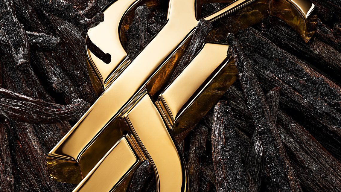Primo piano del logo YSL dorato posizionato tra baccelli di vaniglia, fiori bianchi e accenti dorati scintillanti.