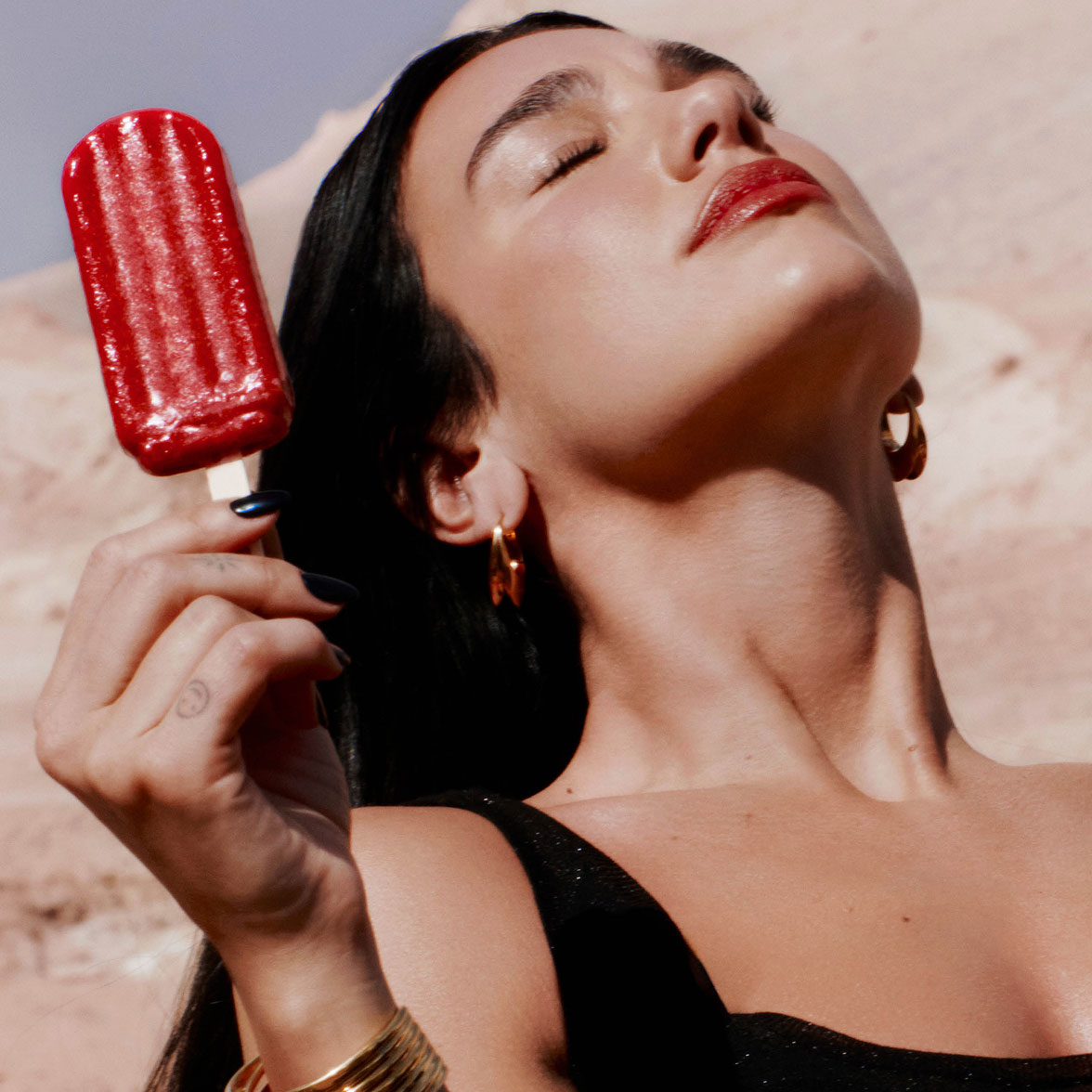 Un primo piano di Dua Lipa con gli occhi chiusi al sole, mentre tiene in mano un ghiacciolo rosso.