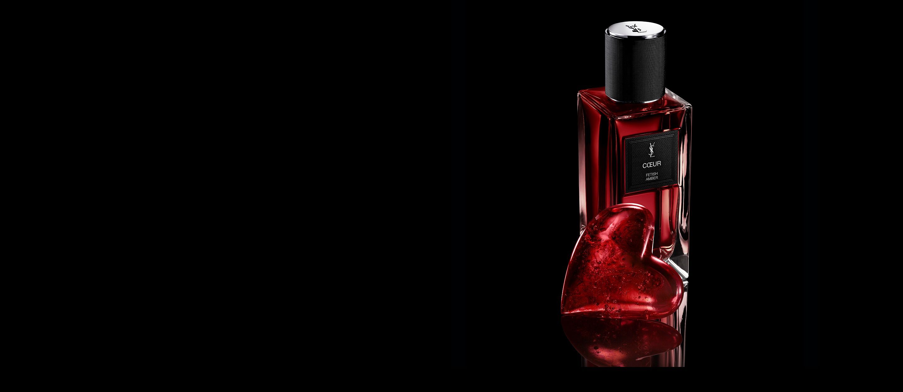 Flacone di CŒUR Eau de Parfum by YSL
accanto a un gioiello a cuore rosso rubino su una
superficie nera riflettente.