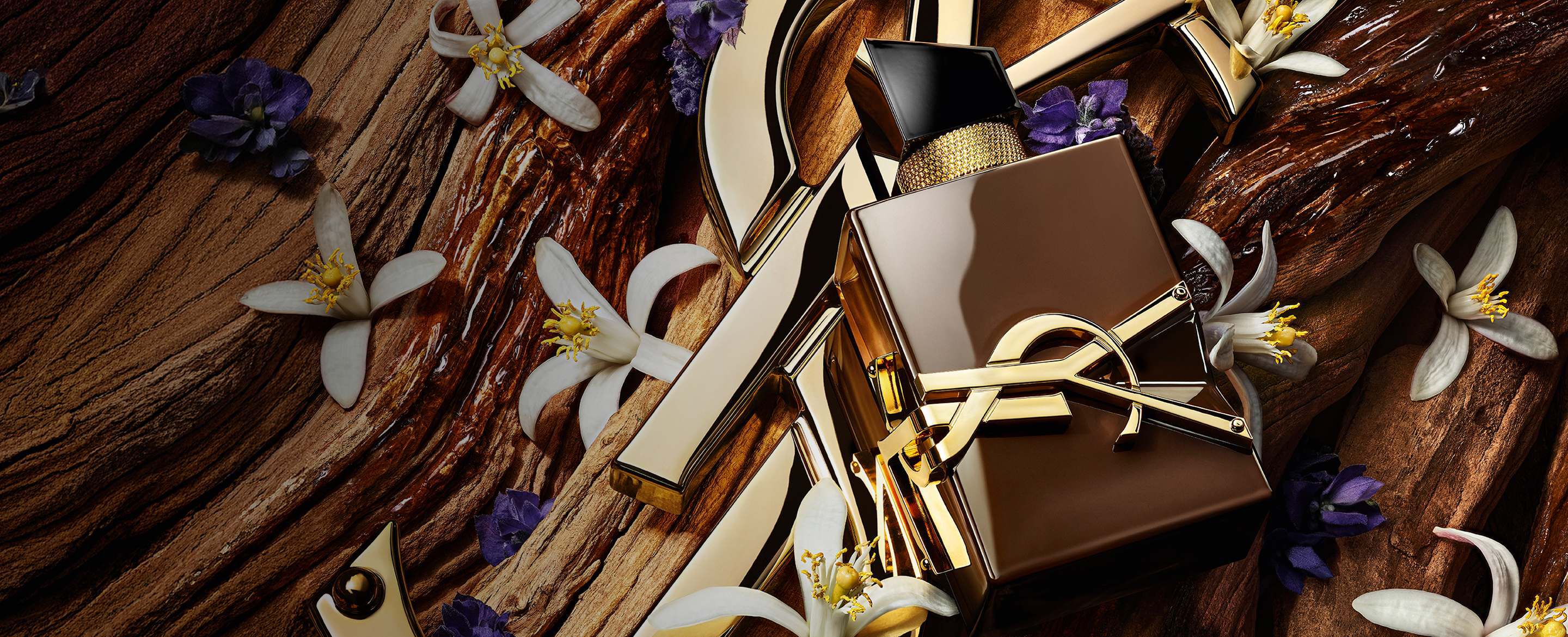 Flacone Libre Santal Couture marrone smaltato che riflette la luce, posato su una grande Cassandre YSL dorata.