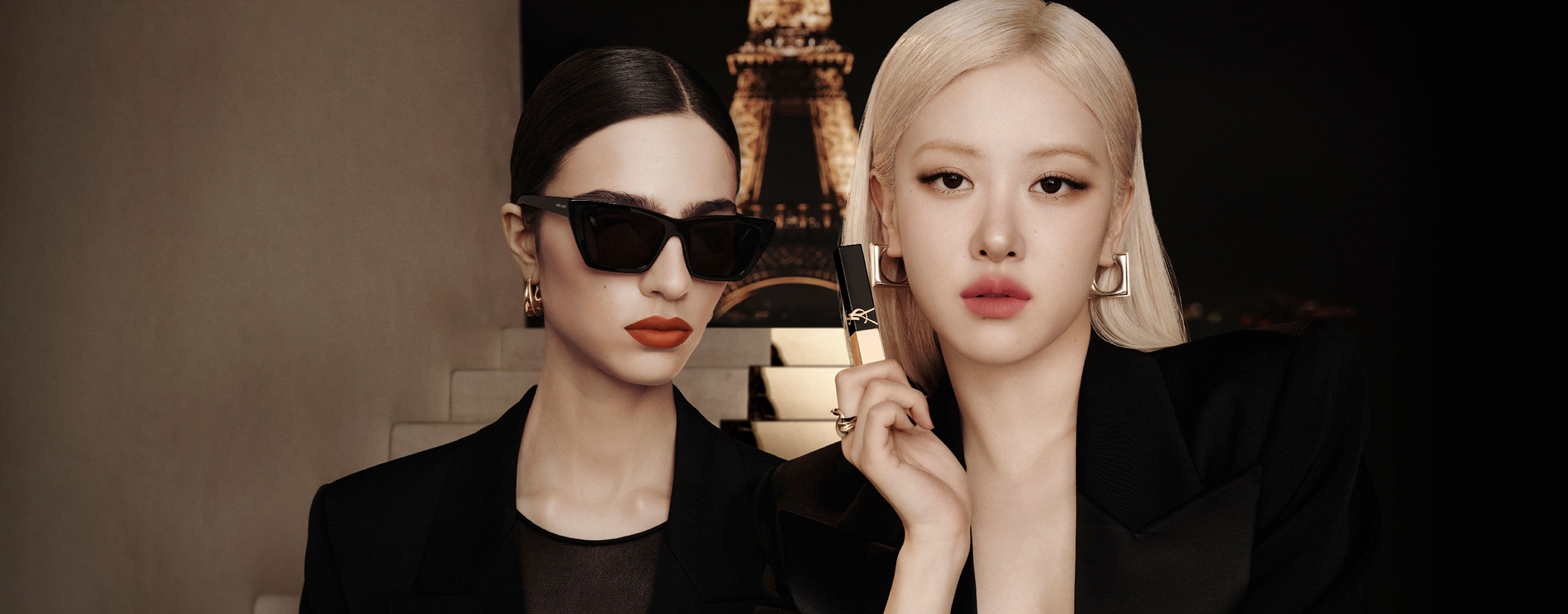 Due modelle in nero davanti alla Torre Eiffel di notte; una tiene un lip product YSL nero‑oro.