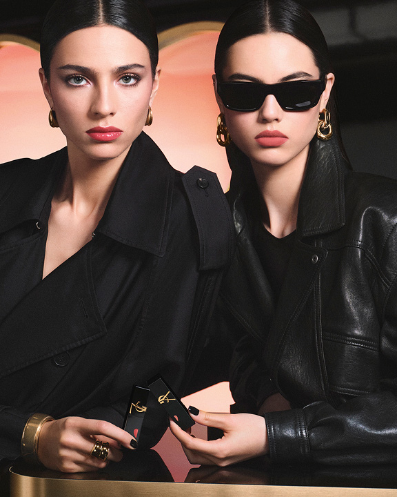 Due modelle chic in trench nero e giacca di pelle reggono rossetti YSL davanti a uno sfondo rosa a forma di cuore, mostrando un look labbra lucide rosse e un effetto matte rosa delicato.