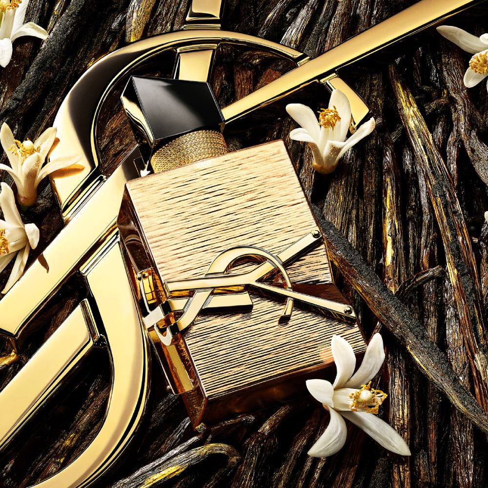 Primo piano della bottiglia dorata Libre Vanille Couture circondata da ingredienti chiave come fiori d'arancio.