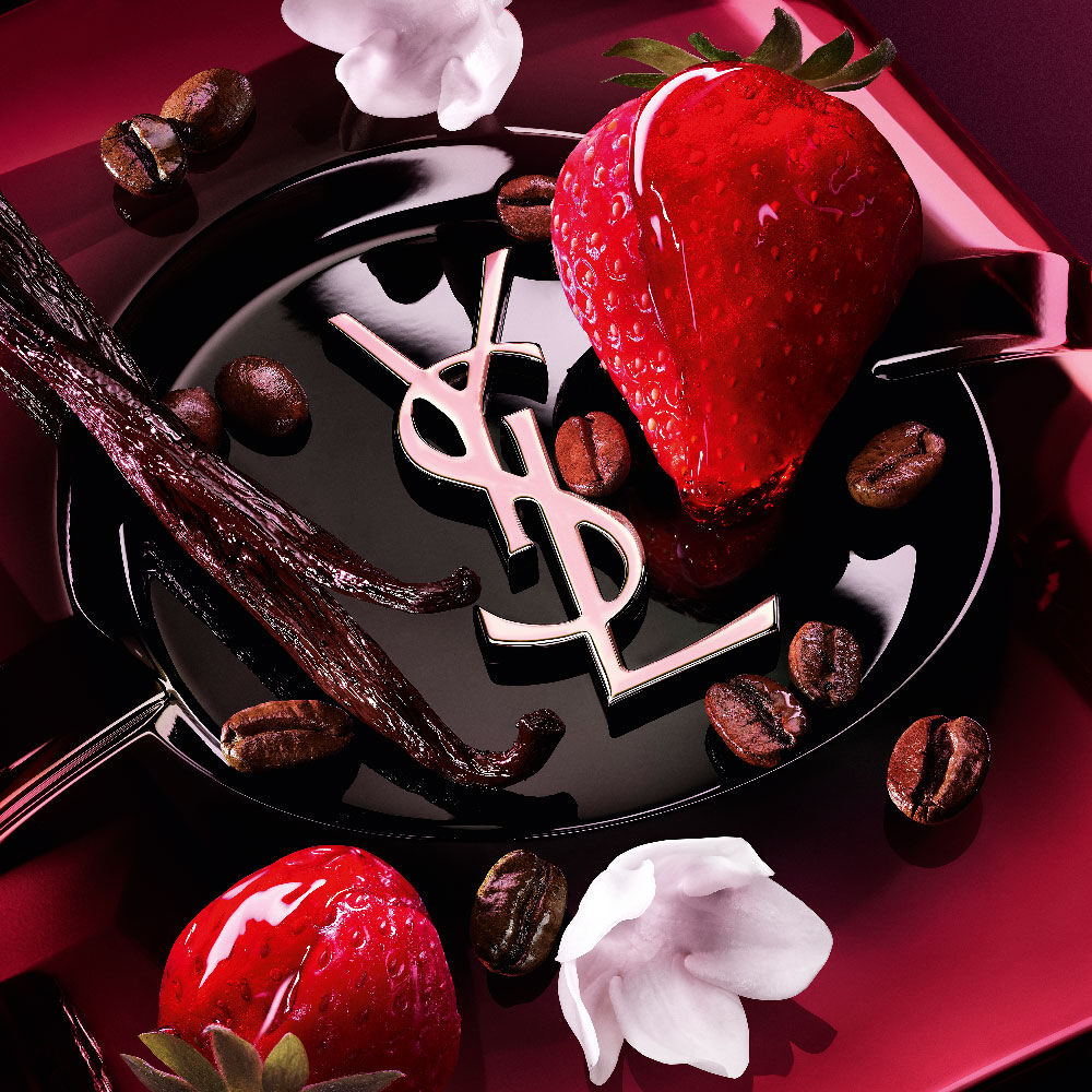 Ripresa
                ravvicinata di fragole fresche, baccelli di vaniglia, fiori di gelsomino e chicchi
                di caffè tostati disposti sulla bottiglia di Black Opium Eau de Parfum Pink Glaze
                con l’iconico cassandre YSL.