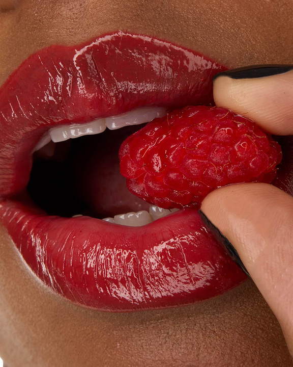 Primo piano delle labbra della modella Zahara, impreziosite dal rossetto YSL LOVESHINE nella tonalità Berry Crush 219, mentre tiene tra i denti una lampone fresco.