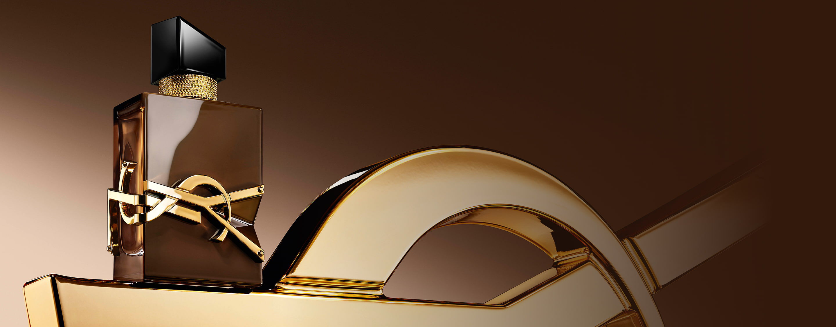 Flacone Libre Santal Couture marrone effetto glassato che riflette la luce, posato su una grande Cassandre YSL dorata.