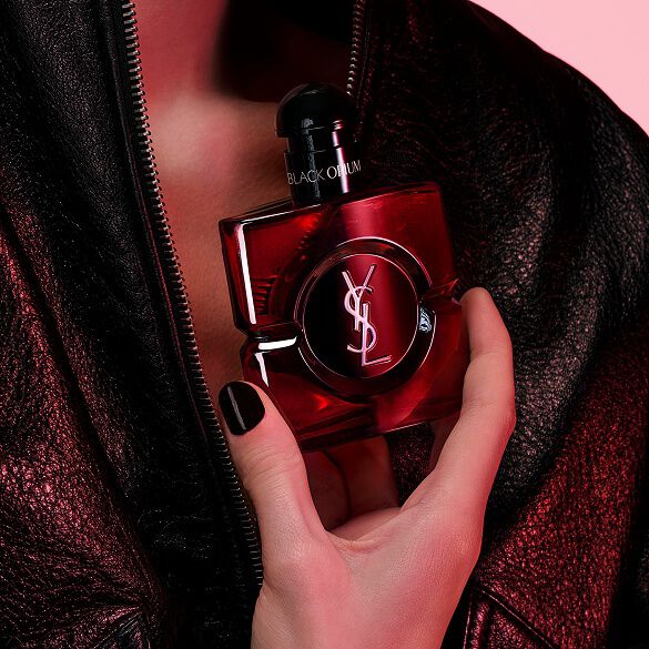 Primo piano di una mano dalle unghie nere che stringe il flacone di Black Opium Eau de Parfum Over Red contro una giacca di pelle nera.