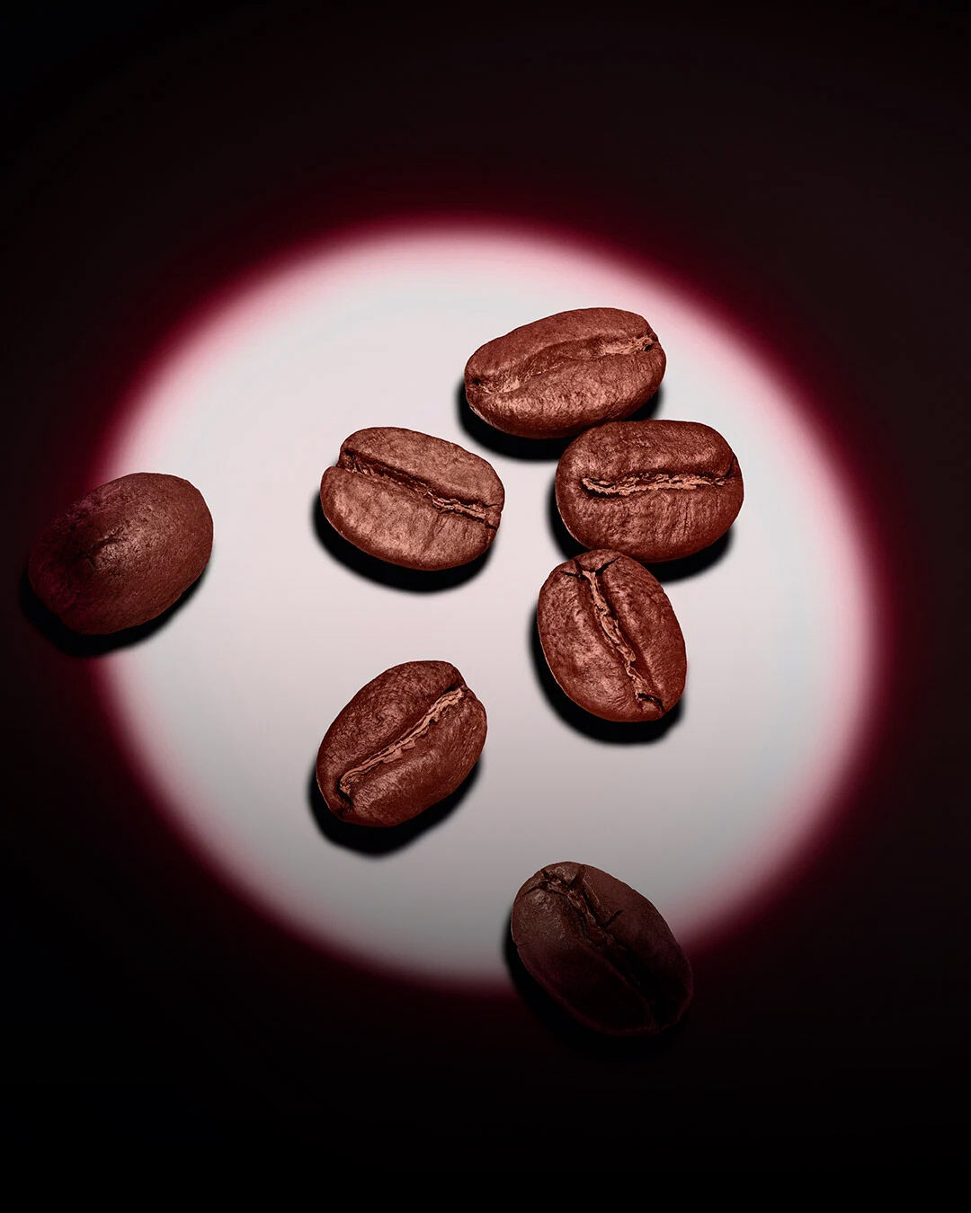 Un gruppo di chicchi di caffè tostati illuminato da un riflettore rosa circolare su uno sfondo scuro. 
