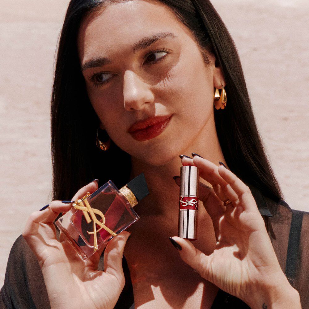 Dua Lipa tiene in mano Libre Eau de Parfum Berry Crush e un YSL Loveshine, con labbra rosse e orecchini d'oro.